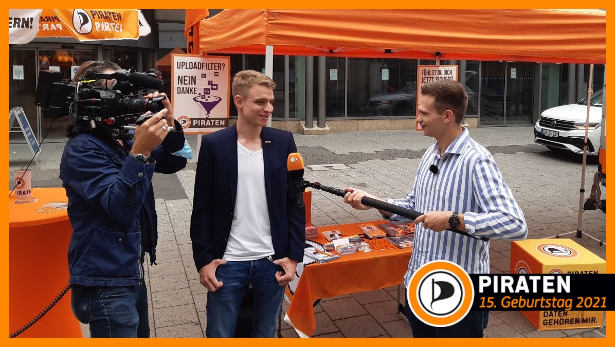 Auf diesem Bild sieht man links einen Kameramann, rechts daneben unseren Bundestagskandidaten Lukas Küffner, und rechts neben ihm Fabian Köstner von der Heute-Show, der Lukas ein Mikrofon entgegen hält. Im Hintergrund sieht man unseren aufgebauten Infostand mit Infomaterial, Plakaten, Schirmen, einem Pavillion und Sitzmöglichkeiten.