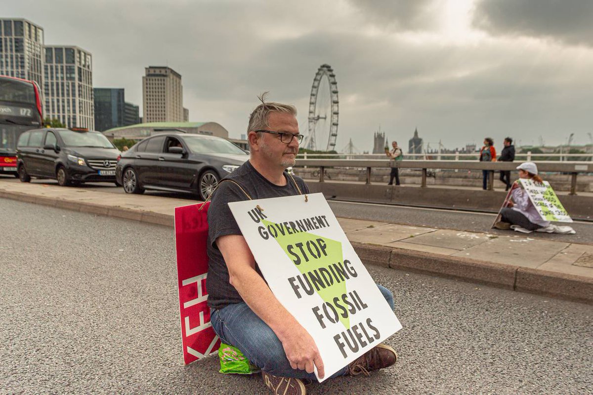 Photo: Extinction Rebellion protest. Rebel with placard sitting in the road “Stop Funding Fossil Fuels” 
Photo credit: Enormis