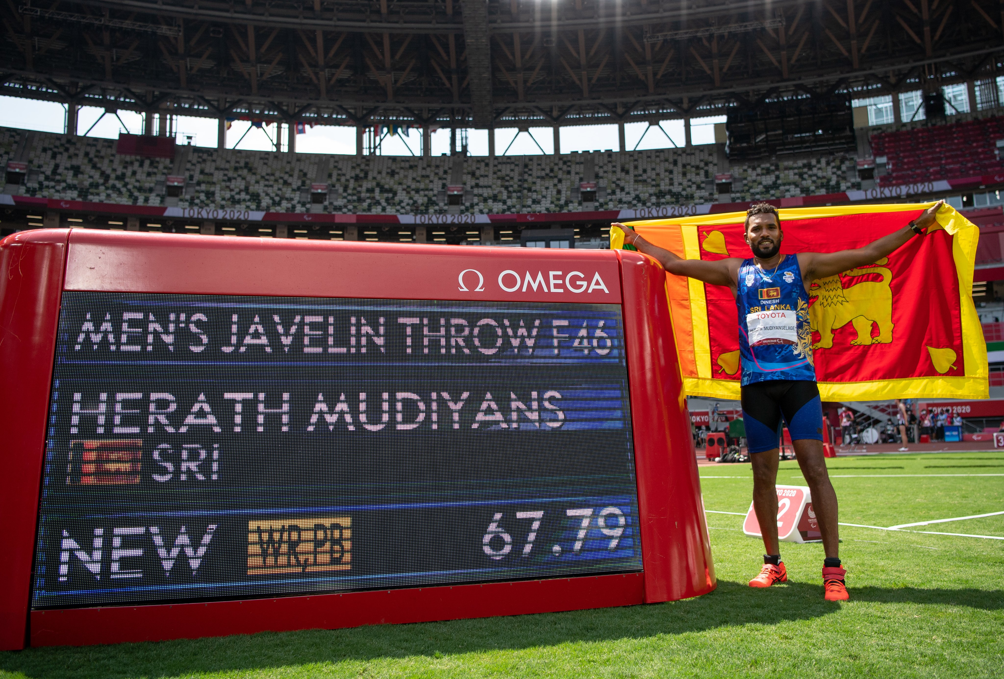Herath Mudiyansalage Dinesh Priyantha celebrates winning gold with the Sri Lankan flag next to the scoreboard.