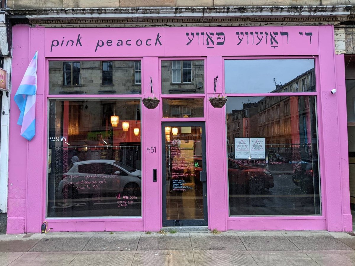 the front of the café: a pink facade with large windows. the sign reads "pink peacock די ראָזעווע פּאַווע". there is a large trans flag and two hanging flower baskets