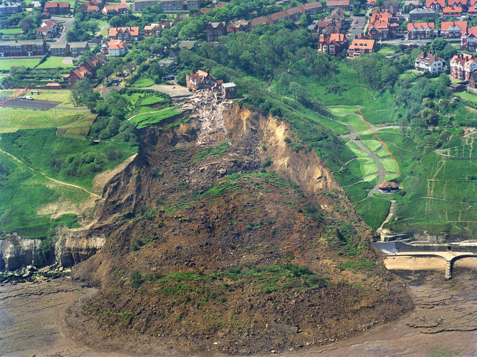 An image of the huge Holbeck Hall landslide. There are buildings and homes at the top of the cliff, which is covered in greenery and the huge slide shows tonnes of earth falling onto the shore below. 