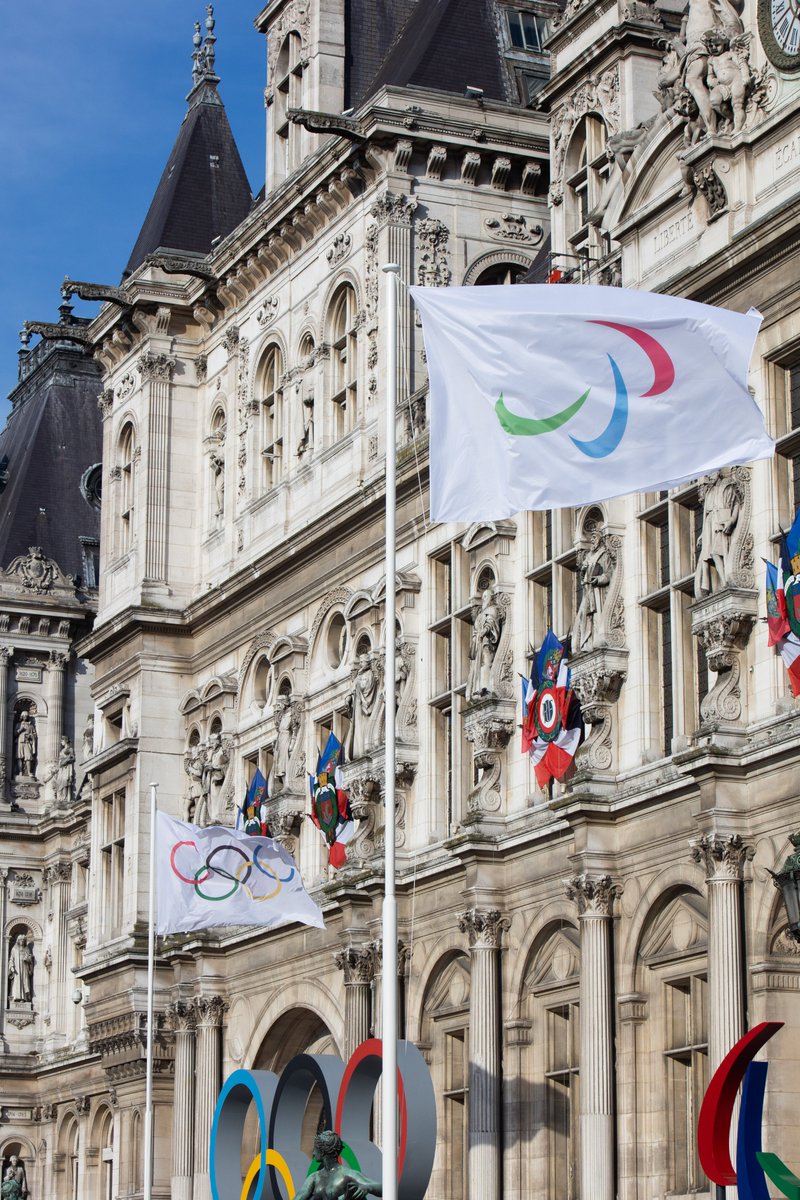Photo du drapeau paralympique qui flotte sur son mât devant l'hôtel de ville de Paris avec le drapeau olympique en fond.