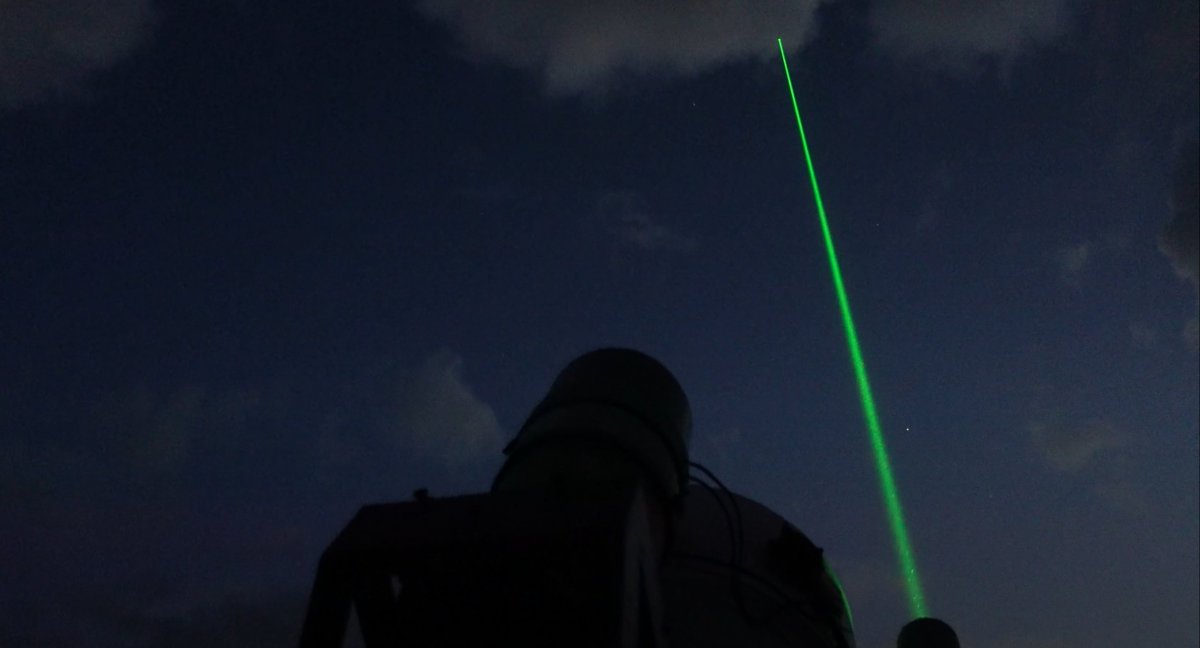 A bright green laser beam shoots from the roof of the facility, which you can just make out in shadow. The sky is dark and you can see stars in the distance, as well as some wispy clouds. 