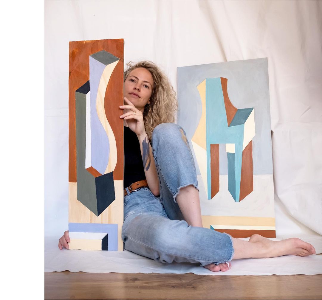 Adriana Jaros sitting on the floor presenting two of her paintings.