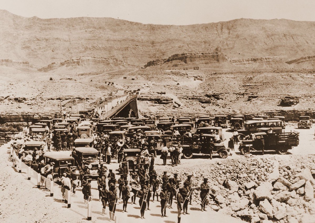 Historic black and white photo of approximately 50 cars and trucks parked on one end of a suspension bridge. On a road in the foreground is a marching band holding their instruments. A desert landscape is visible in the background.