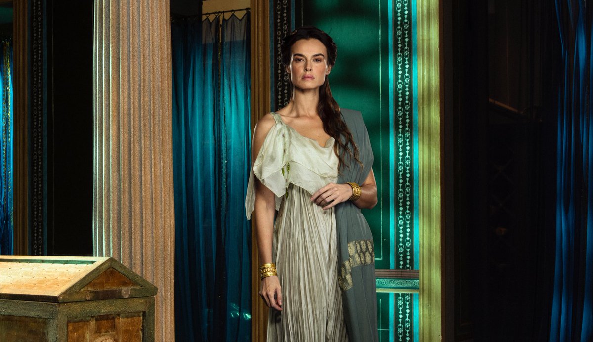 Kasia Smutniak as Livia in a promo shot for the upcoming limited series Dominia. Livia stands in a room with dark teal curtains and wall fresco detail while next to what looks like a sarcophagus with a column in the background. Source: Evening Standard