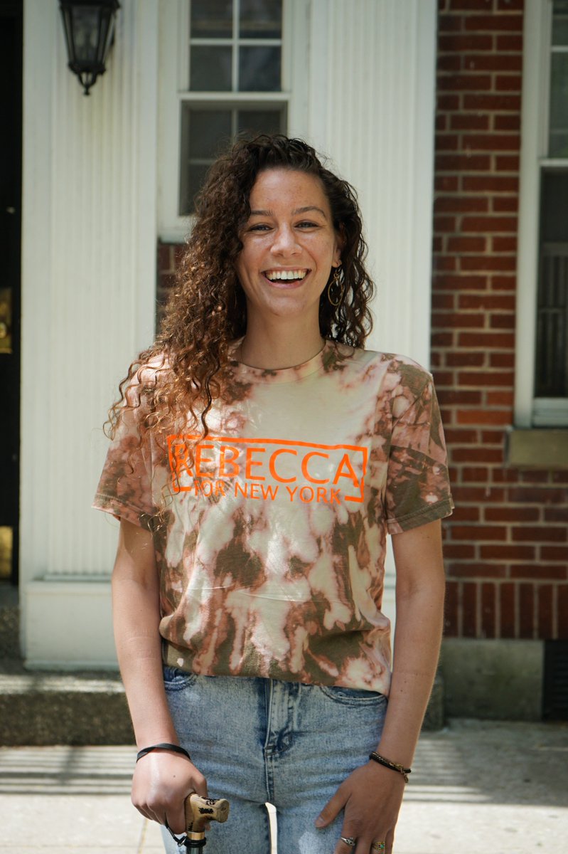 Rebecca, a white woman with curly hair, is smiling and standing in front of a brick building wearing an orange tye-dyed Rebecca For New York t-shirt.