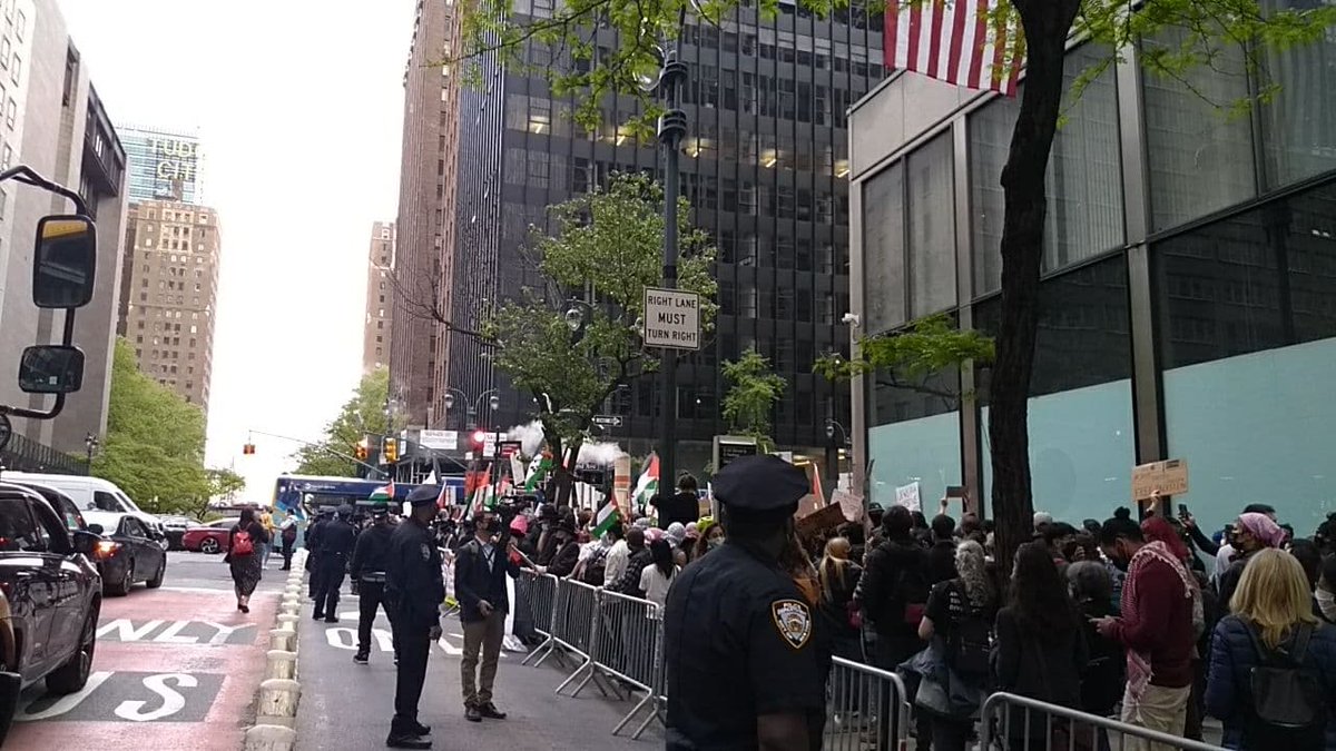 Police officers stand in the street, next to metal barricades with protesters on the other side. Some protesters are waving flags and wearing keffiyeh.