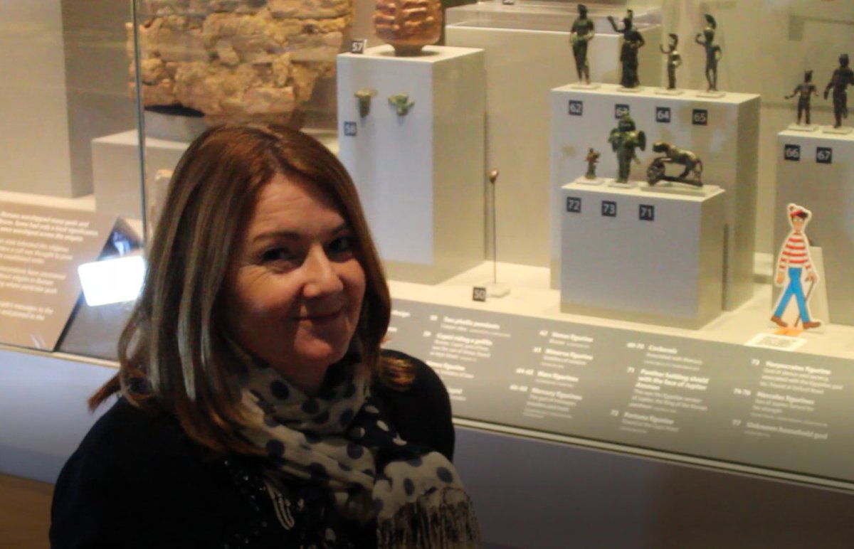A photograph of a smiling white woman with shoulder length brown hair. She is next to a display case at Colchester Castle, containing an assortment of small bronze figures. A Where's Wally cartoon is at the front of the case