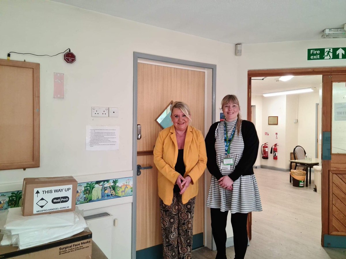 Two NHS staff standing in Grove Road Health Centre, Wrexham, by a stack of boxes of PPE.