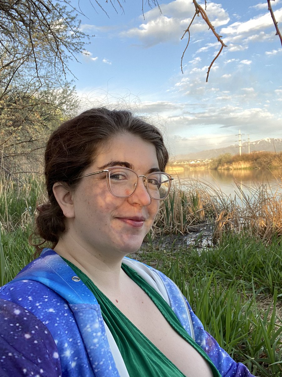 Smiling selfie of white femme with glasses, in front of river, with dappled light on her face