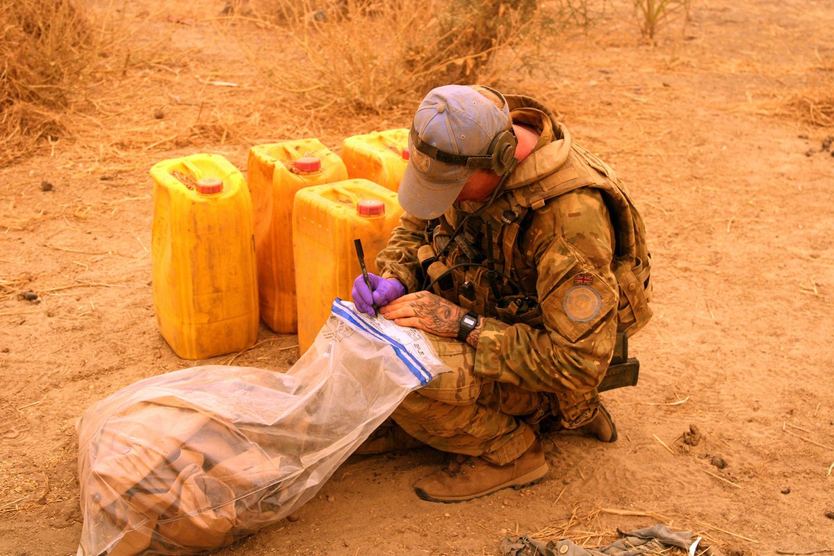 A soldier in a blue UN hat crouches next to yellow fuel containers and writes on a plastic evidence bag of seized material.