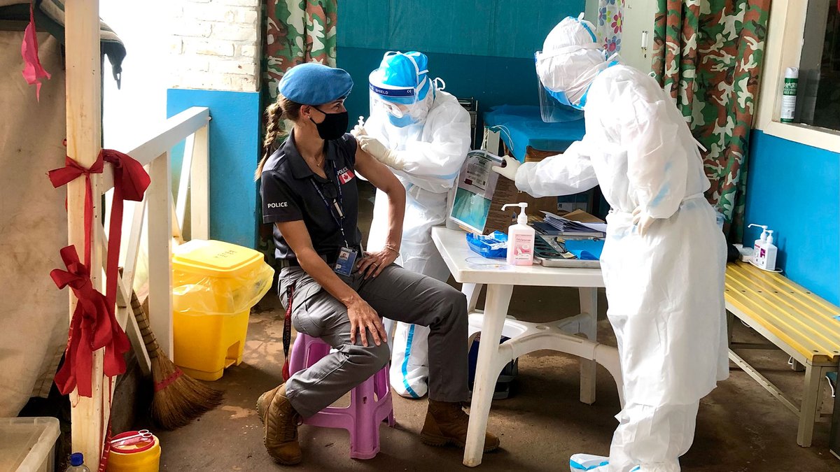 A Canadian police officer is sitting at a table with two medical personnel standing beside her administering the COVID vaccine in the Democratic Republic of the Congo.