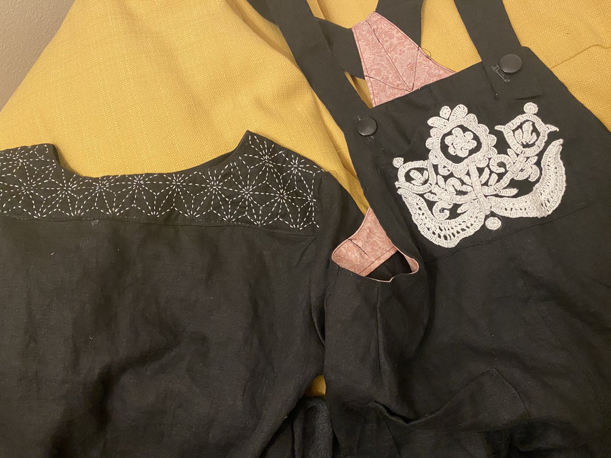 Two pieces of black clothing are laid on a yellow background, cropped to include only the top halves. The left piece is the back of a dress whose yoke is embroidered with a white geometric pattern. The right piece is a pair of overalls with a white rustic folk embroidery done with yarn on the large front pocket.