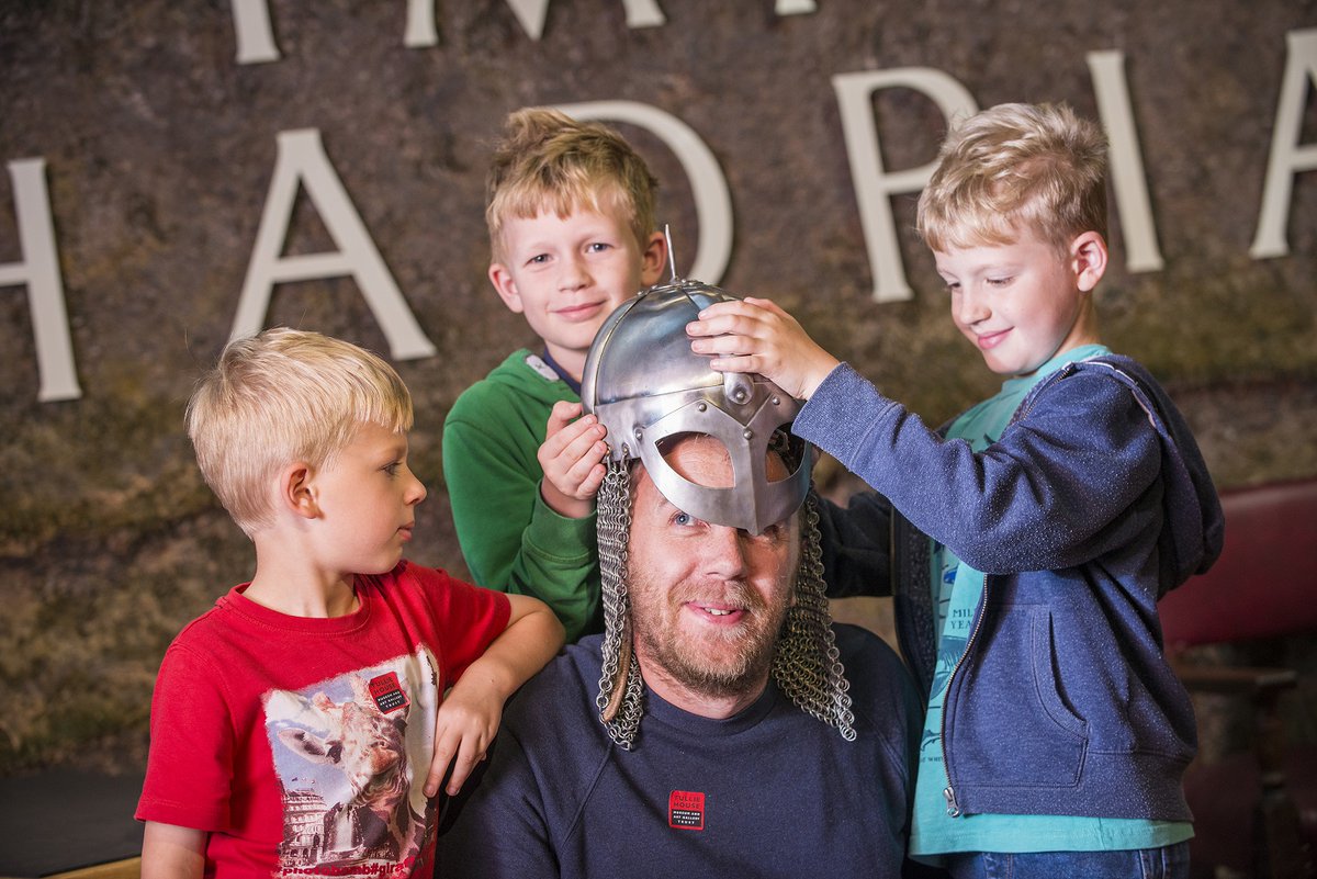 Three young boys surround their father who is kneeling and place an old helmet on his head.