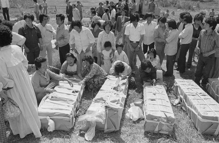 A crowd gathers around four covered caskets. Several women kneel before them weeping. One older woman looks off with a faraway look in her eye. Behind them stand several men and some women and children, all in various expressions of mourning or anger