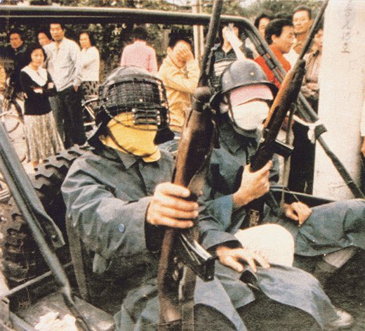 Two civilian militia members with M1 Carbines. Their faces are obscured by helmets, kundo masks, and cloth. They are both wearing long trench coats. Behind them a group of people look on