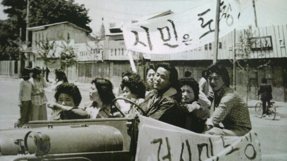 Young students in what appears to be a liberated military vehicle. They look placid, carrying banners somewhat obscured by the camera angle. Behind them a group of young women are engaged in conversation