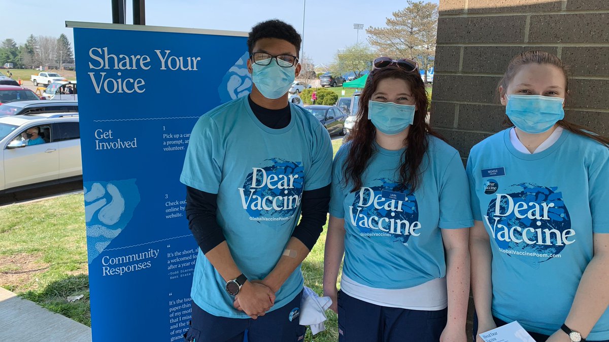 A photo of three people wearing blue "Dear Vaccine" t-shirts standing next to a blue banner that reads "Share Your Voice" at a mass vaccination site at the Kent State University Field House.
