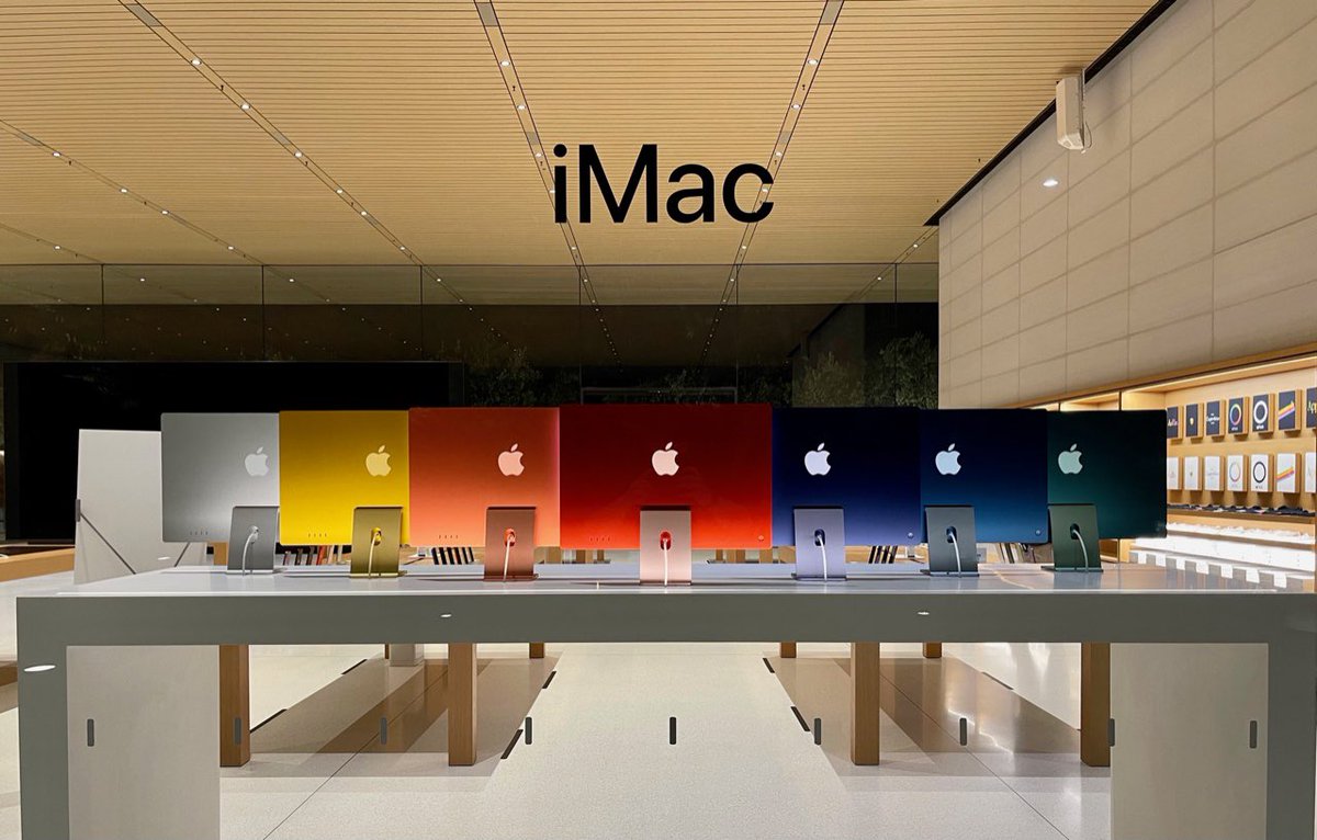 Through the glass window of an Apple Store, seven iMacs in silver, yellow, orange, red, purple, blue and green are seen sitting on a white table. “iMac” is written on the glass above the display. It is night time and the store behind the iMacs is empty.