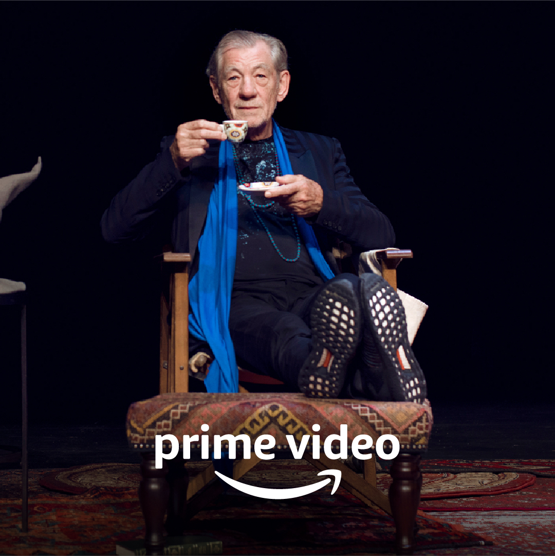 A man with grey hair, wearing black with a blue scarf and necklaces, drinks from a patterned espresso cup. Prime Video logo.