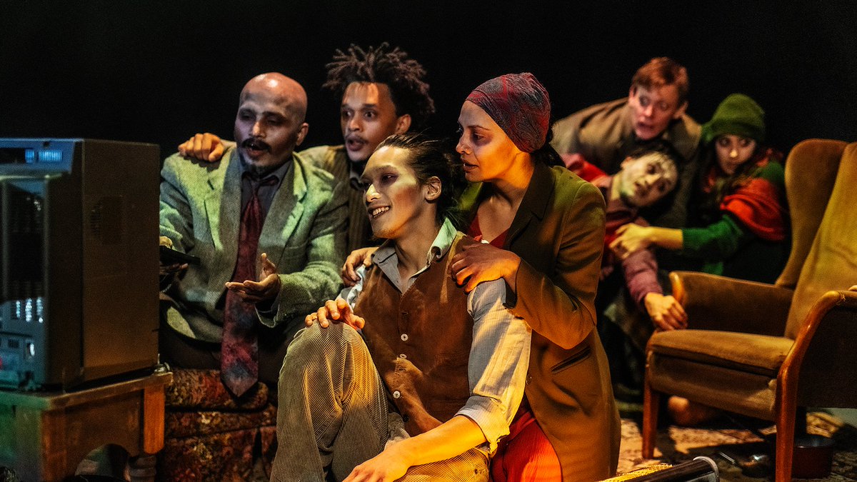 Four people in motley clothes with white make-up and reddened eyes look at an old box TV.