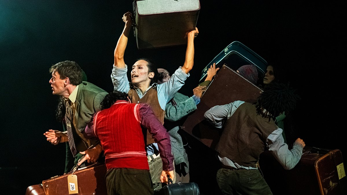 A group of people in colourful clothes move around each other holding old fashioned suitcases and trunks, looking concerned.