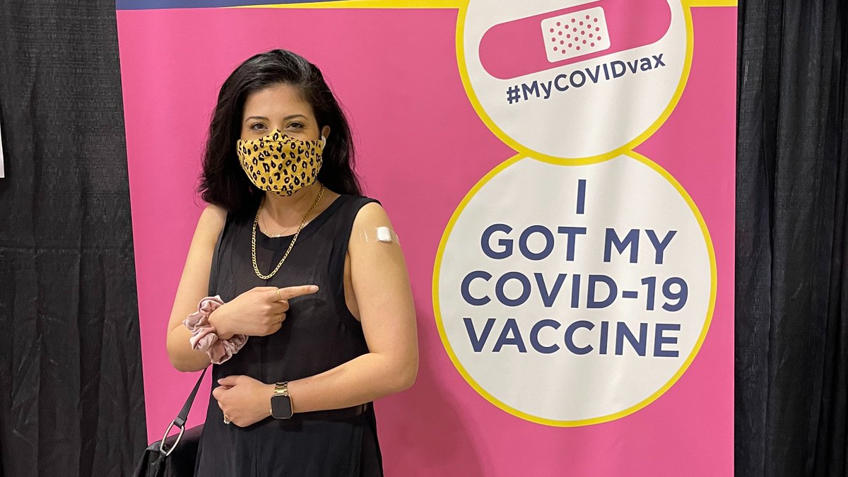 Samantha with a leopard-print mask and smiling eyes pointing to her bandaged white arm standing in front of a backdrop that says "I got my COVID-19 vaccine"