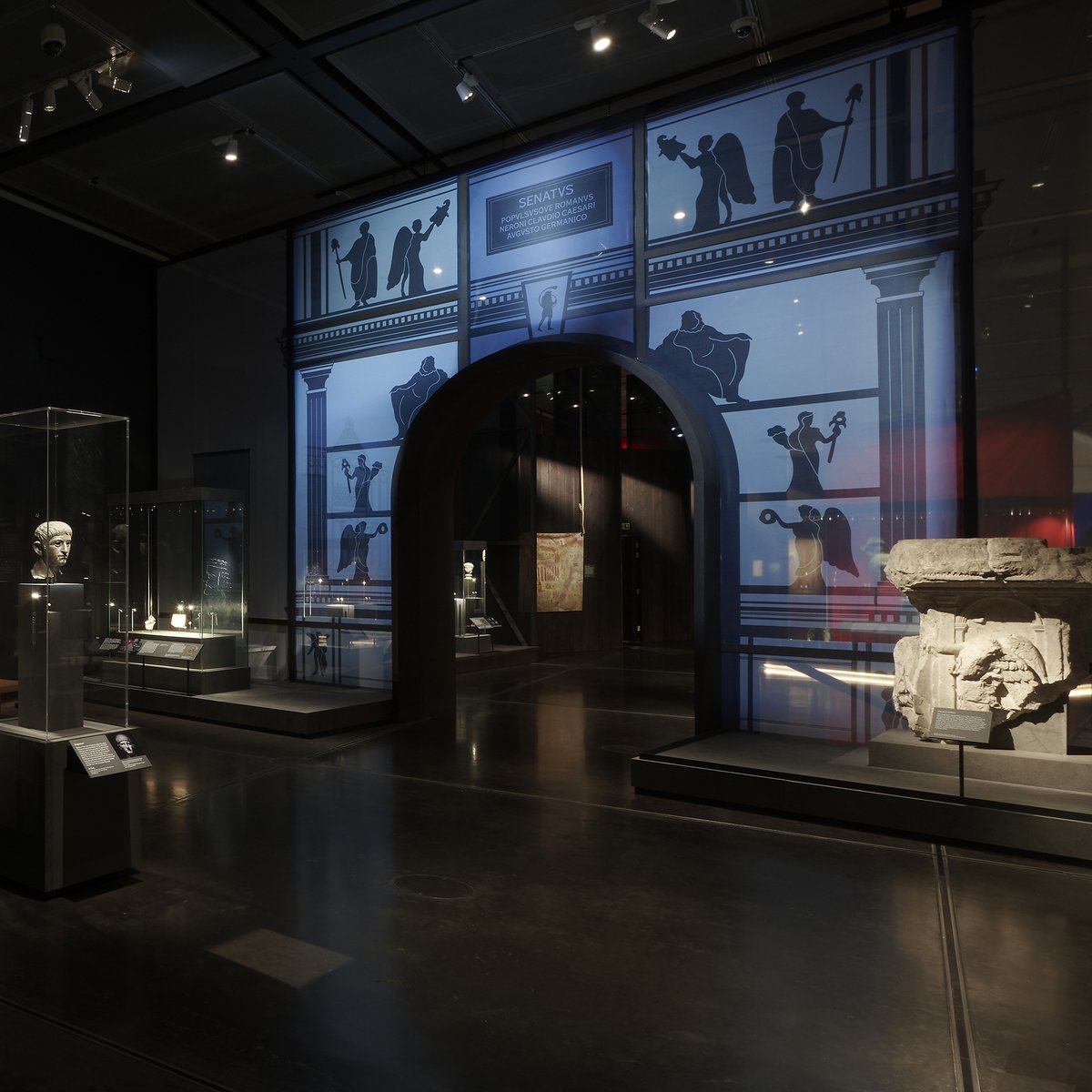 Inside the exhibition - a large blue triumphal arch divides the space, and there is an architectural fragment on the right and a bronze bust of Nero on the right. 