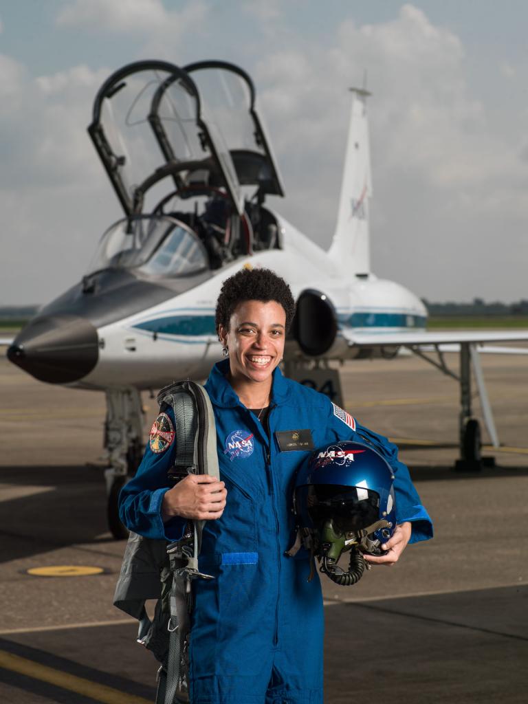 Jessica Watkins posing in a blue flight suit holding her training helmet and flying gear as she stands in front of a NASA T-38 training aircraft.