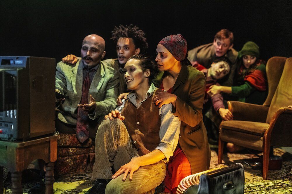 Image of performers dressed up in semi-formal clothing sat around a television with chairs, rug and heater