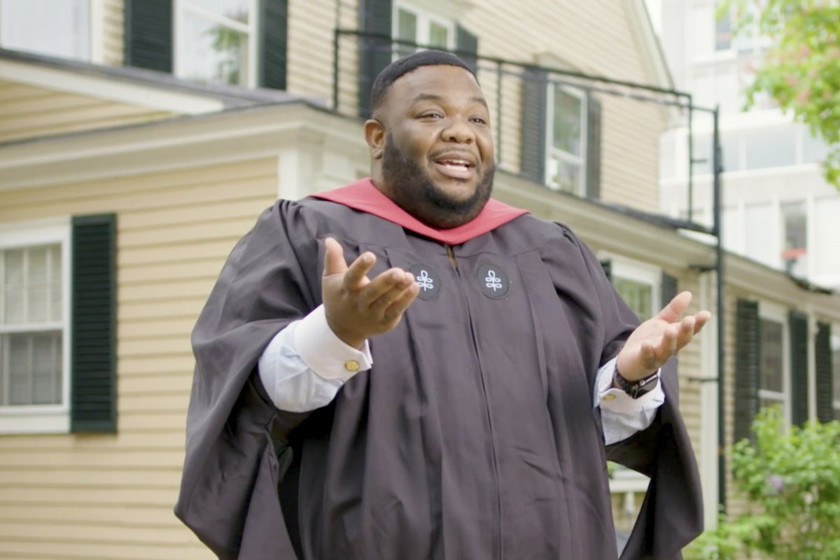 A graduate wearing a graduation gown and a crimson hood stands outside 