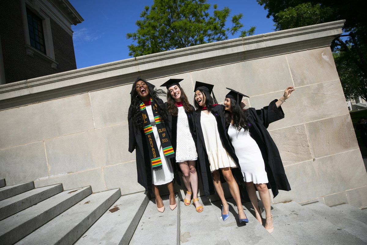 Four graduates wearing gowns over white dresses pose on the steps of Widener Library 