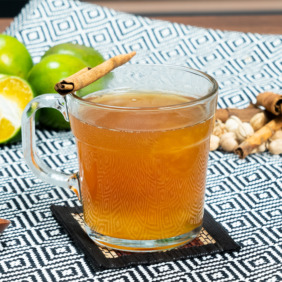 A glass mug of warm apple cider or tea garnished with a cinnamon stick, accompanied by green apples and additional spices on a patterned cloth.