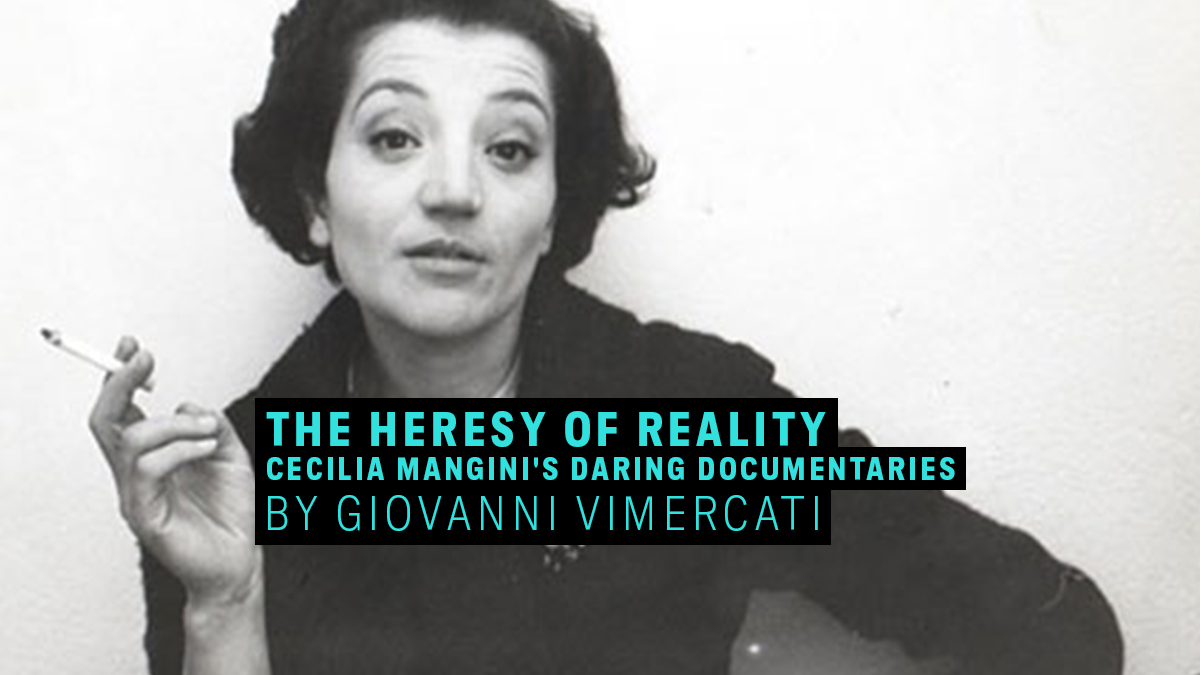 A woman with dark hair and wearing a dark shirt holds a cigarette while looking directly at the camera.