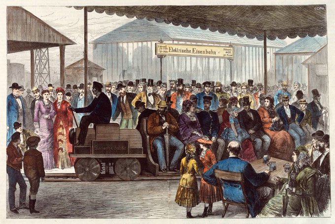 painting of the unveiling of the first electric train in 1879. A large crowd surrounds the Siemens and Halske brown train which sits on a track.