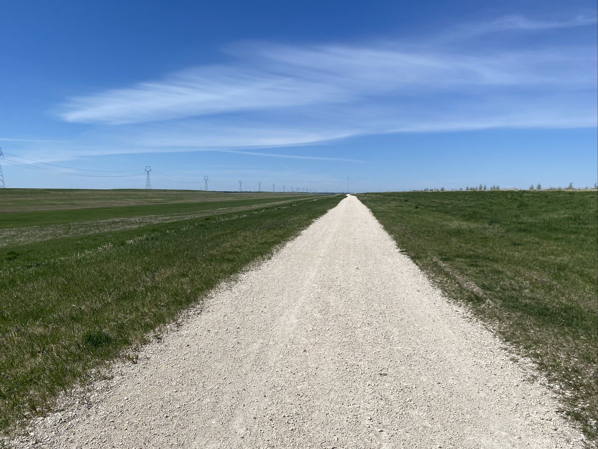 Blue sky with wispy white clouds, beige gravel trail, bordered by green grass on either side. The land is flat.