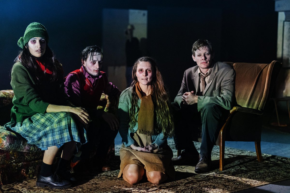 Performers sit in a living room as a family, looking directly at the camera
