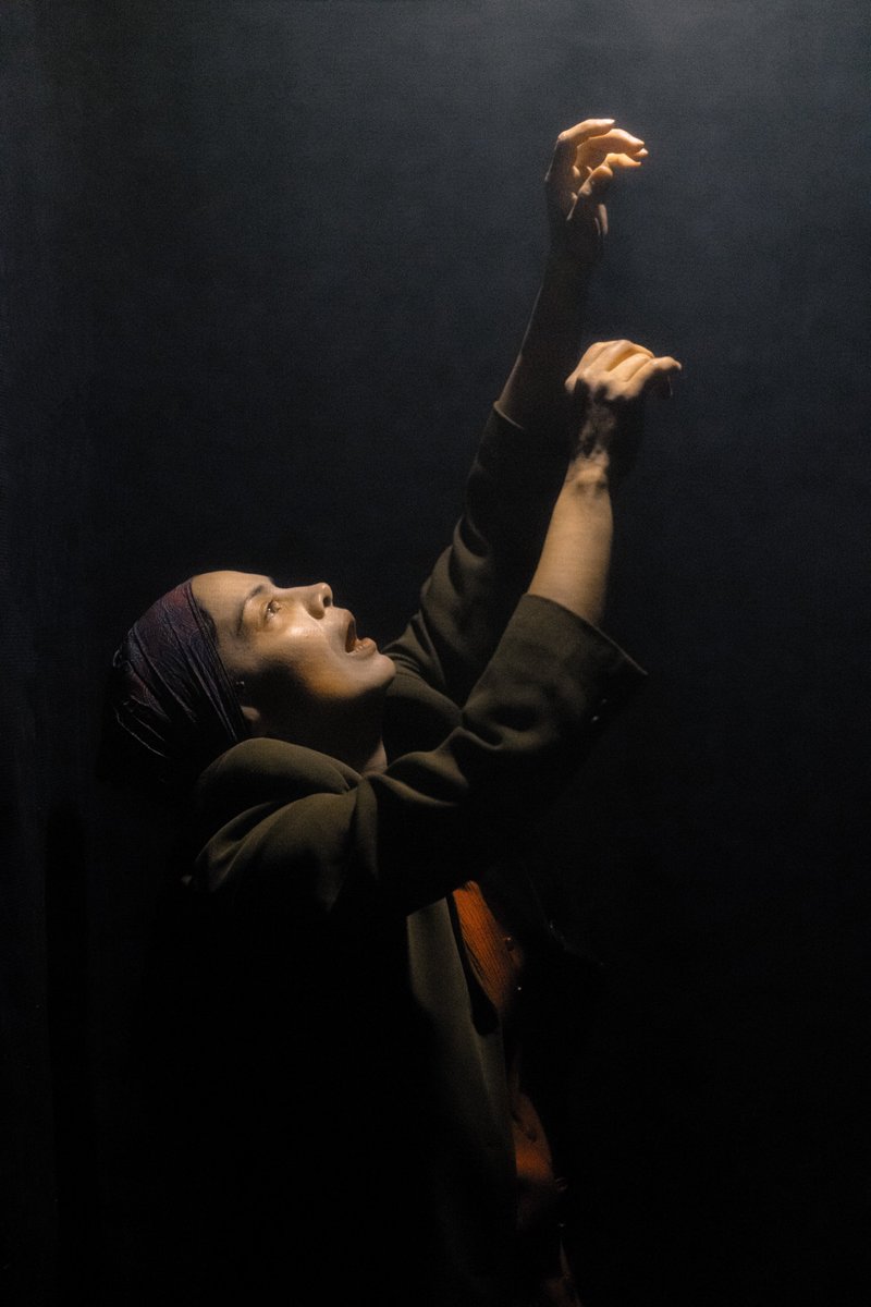 A performer reaches up in the darkness towards a light