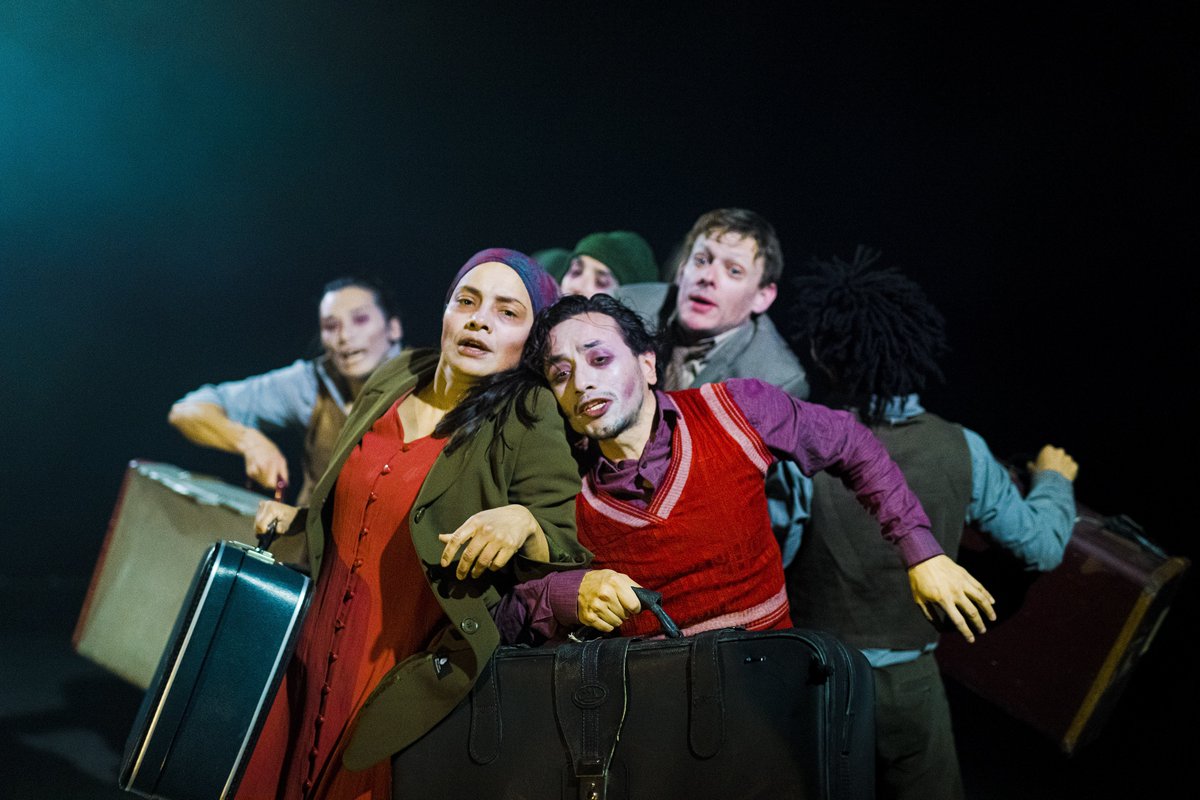 Performers carrying suitcases rush past each other in a crowd, pushing against each other