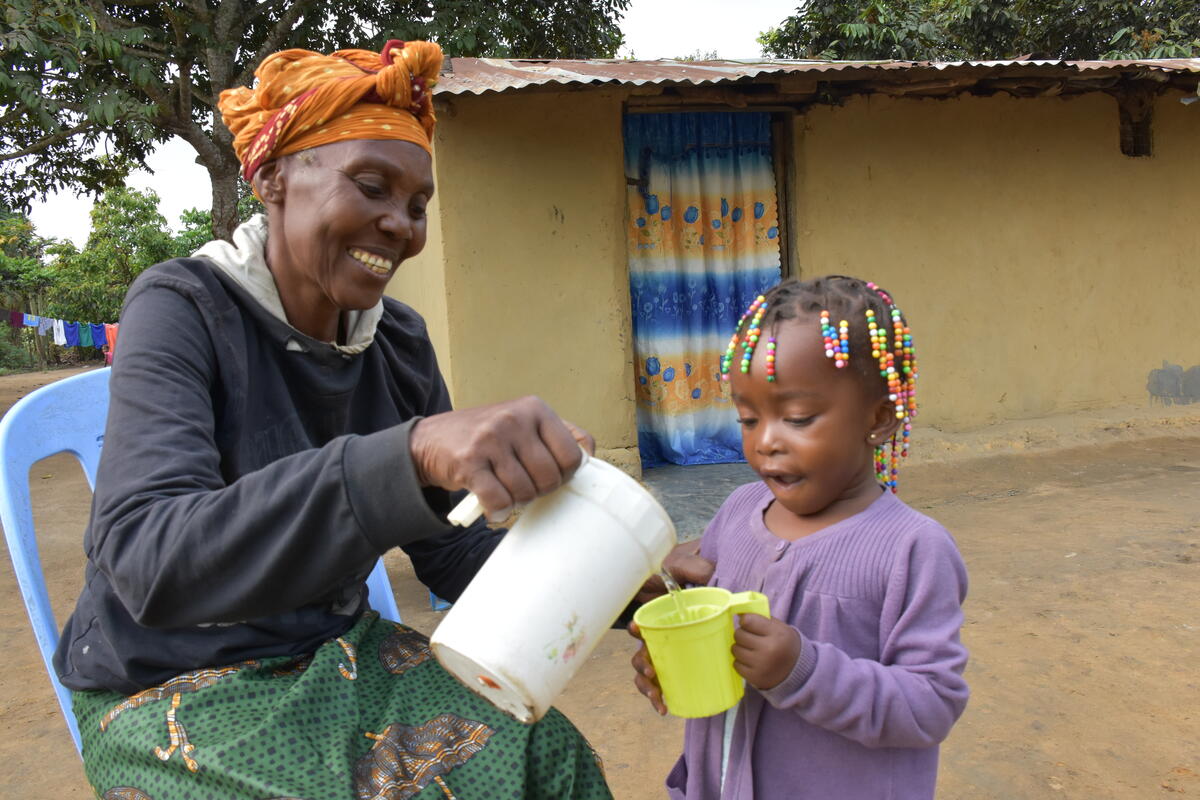 Brigitte is sitting in a chair outside, pouring water from a white pitcher to Jessica's small yellow cup.