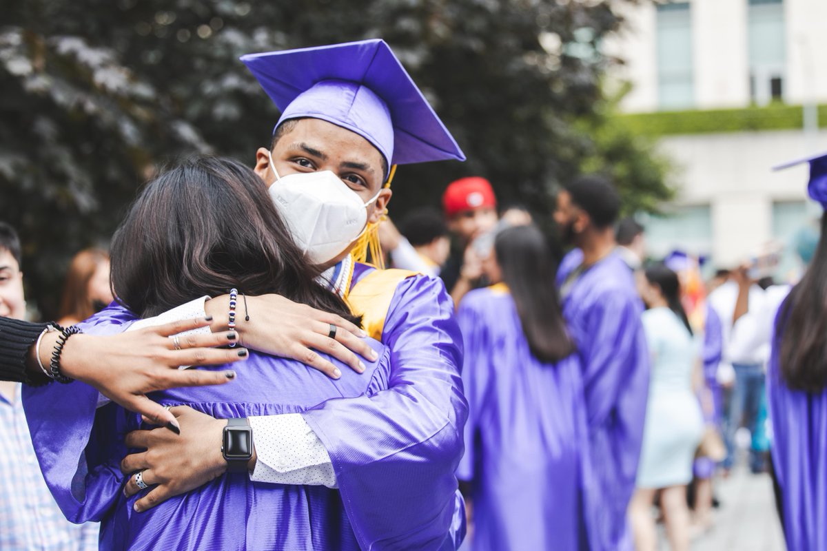 Two people wearing masks and purple graduation gowns embrace in a hug on Lincoln Center’s Hearst Plaza. 