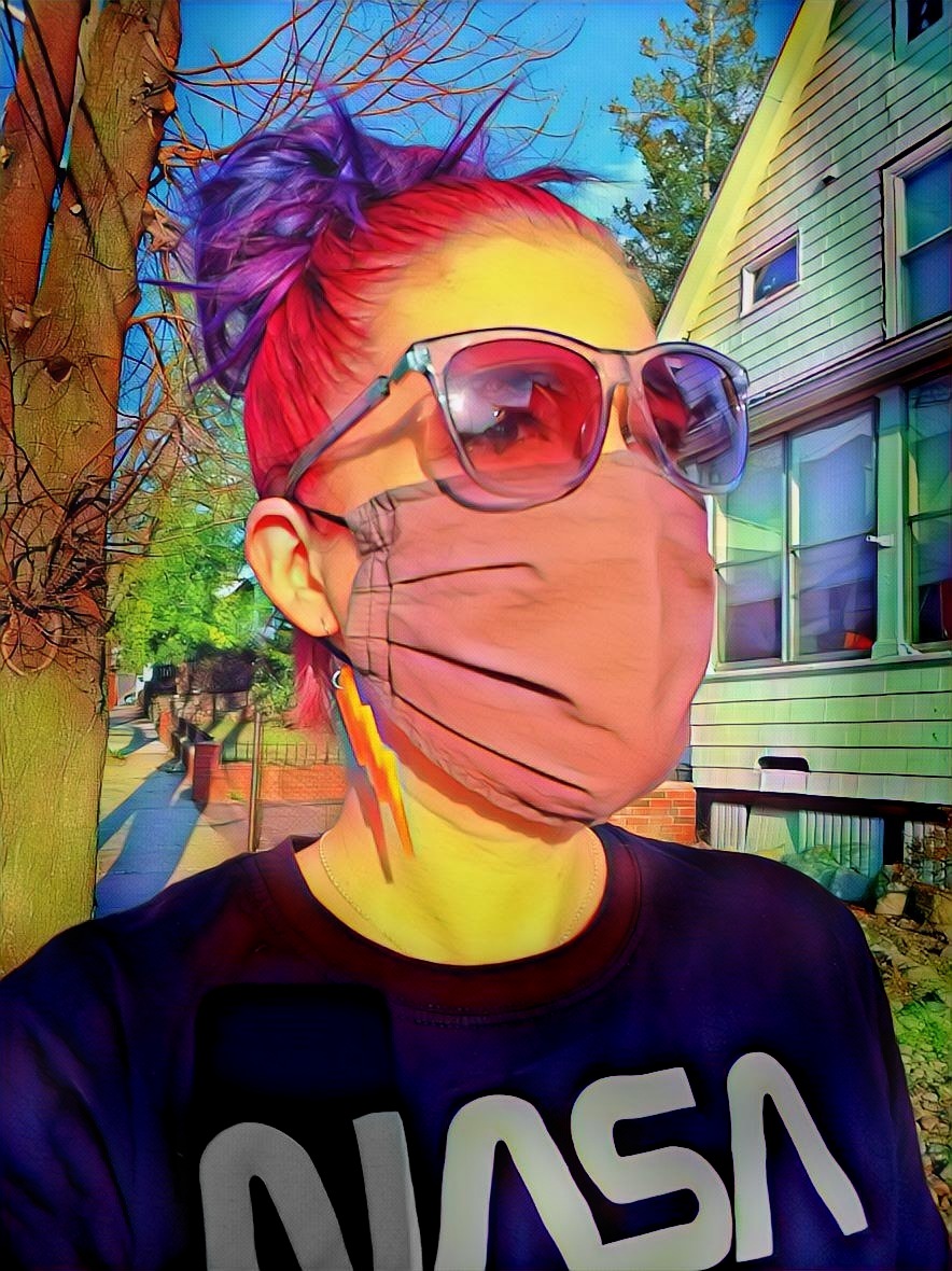 A purple-haired editor wearing purple sunglasses, a gray face mask, and NASA t-shirt walks outside and looks away from the camera.