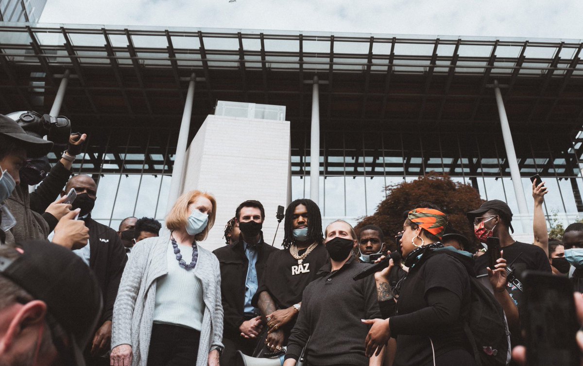 Nikkita Oliver delivers demands to Mayor Durkan. Durkan is in grey clothing while many around her, including Nikkita, wear mostly black. Several microphones and camera phones in front of Seattle City Hall.