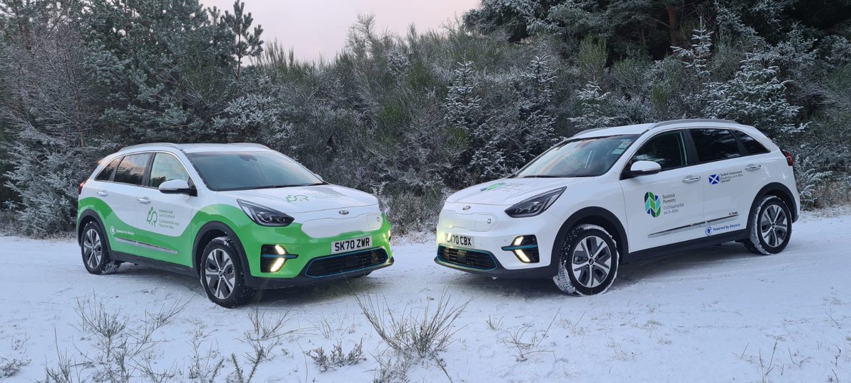 Two electric cars parked on a forest road covered in snow with trees behind