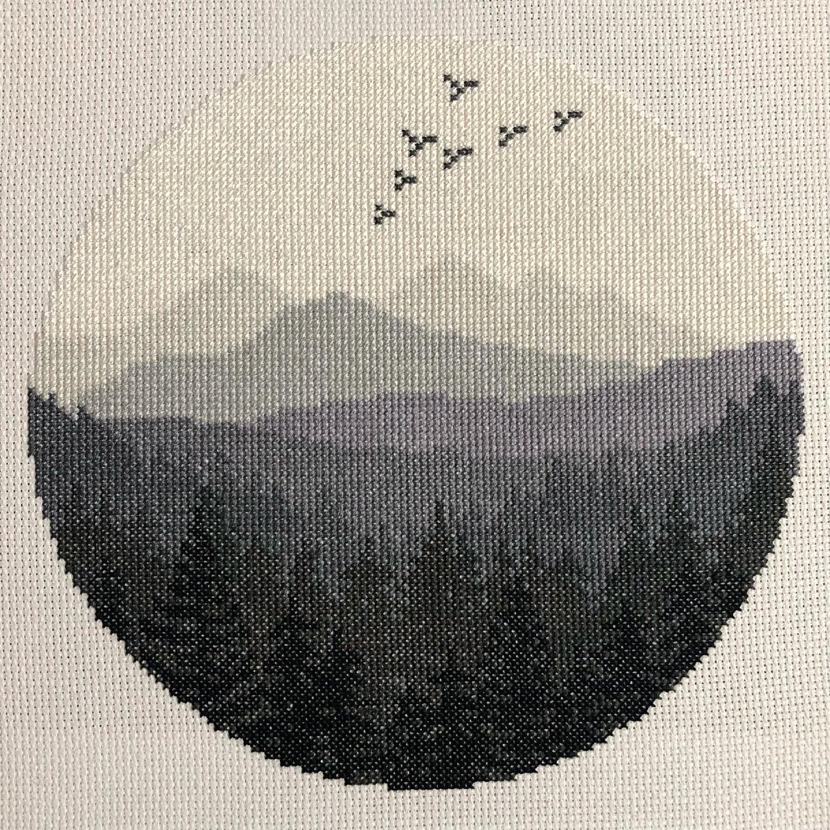 A large circular cross stitch of foggy mountains, with trees silhouetted in the foreground, mountains in the middle, and a big swatch of white sky with a few birds at the top. It’s done all in shades of gray and white, with the darkest colors at the bottom fading gradually toward the top.