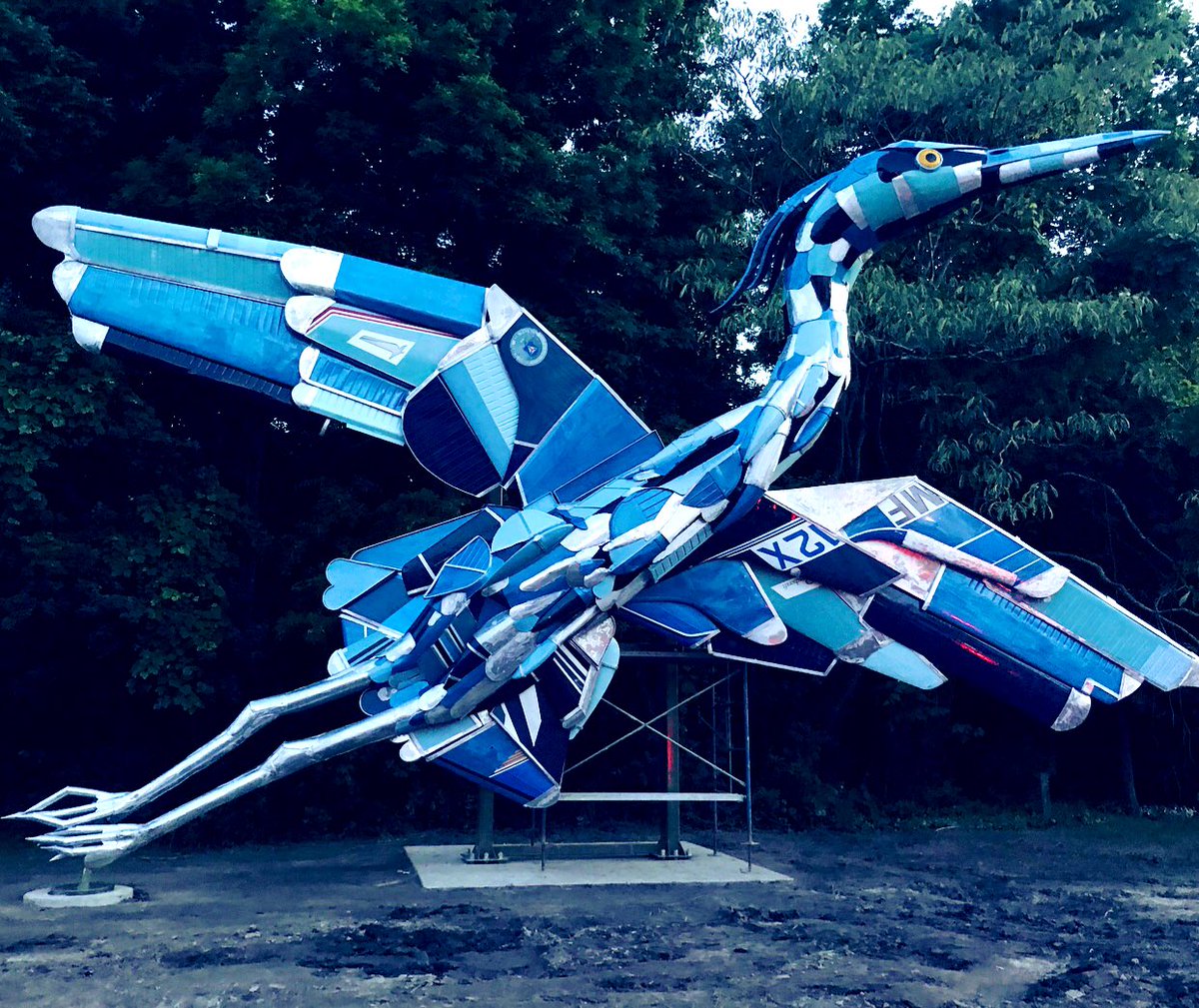 This is a blue heron sculpture that spans over 40 foot wide. It is created from decommissioned airplane parts in a bright blue and white color. It reaches 24 foot high and stands before trees in the background with legs almost touching the ground.