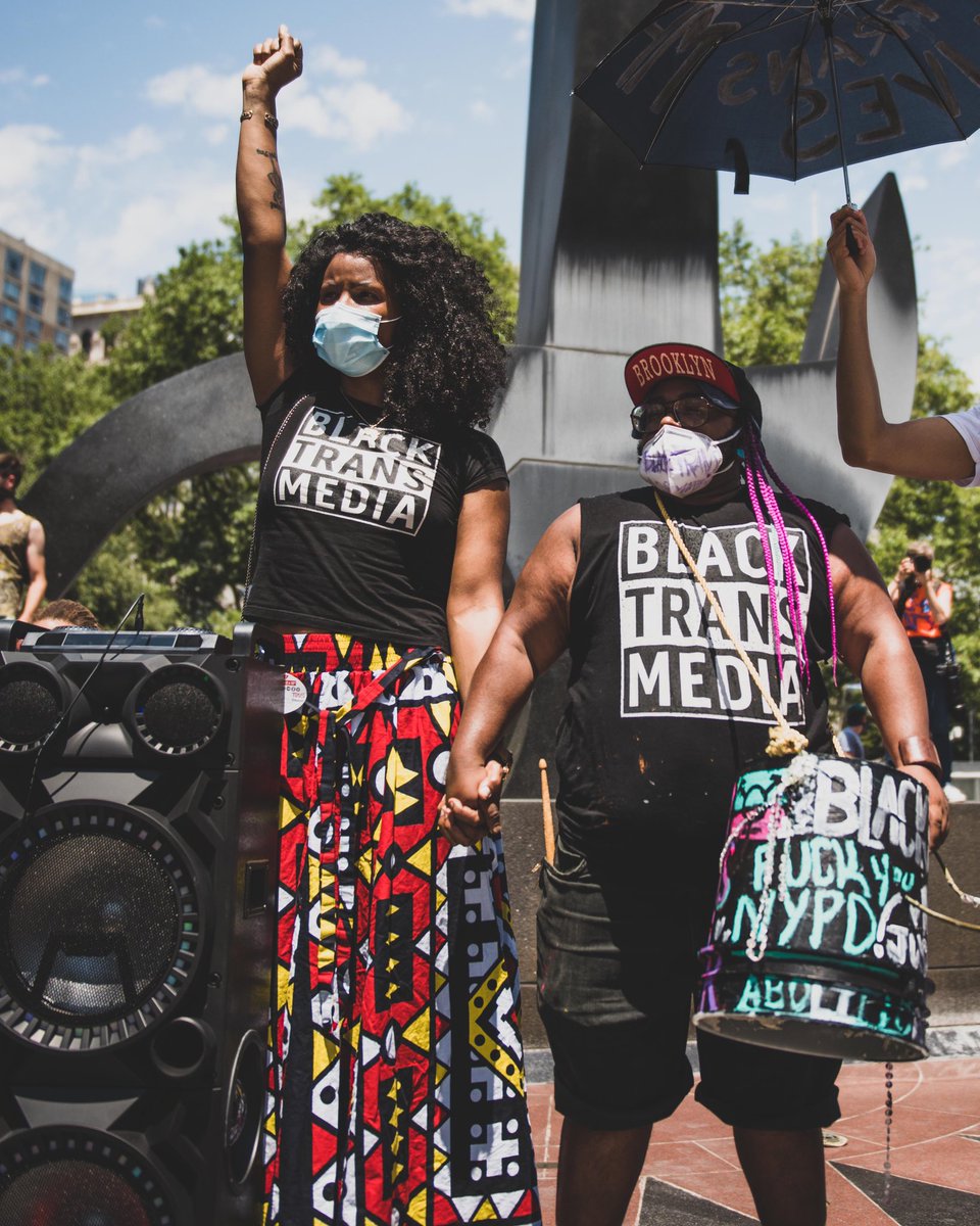 Two black people stand outside, behind a massive speaker. Both people wear shirts reading “BLACK TRANS MEDIA.” On the left, the black person with shoulder-length curly hair holds up their fist. They hold hands with a black person wearing a “Brooklyn” hat and carrying a large drum. The drum is covered in graffiti, and what can be seen reads “FUCK YOU NYPD.”