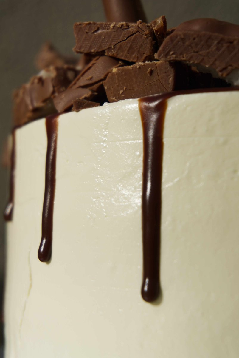 A white birthday cake, with chunks of different chocolate bars at the top and chocolate dripping around the sides, catching the sunlight.
