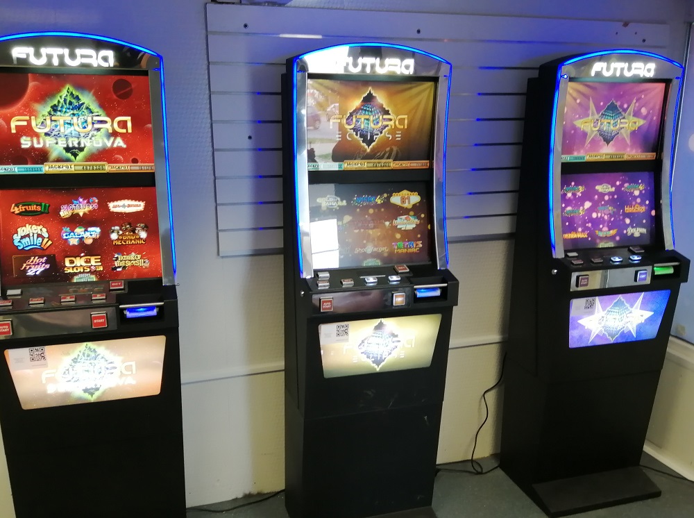 Trzy czynne automaty do gier w lokalu na tle jasnej ściany z roletą.